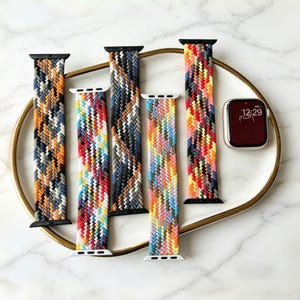 Puede incluir: Correas de reloj trenzadas en varios colores, incluyendo naranja, rojo, azul y amarillo, se muestran en un plato blanco. Las correas tienen conectores negros o blancos. También hay un reloj inteligente con una pantalla roja que muestra la hora.