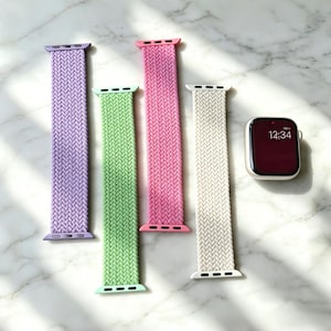 May include: Four braided watch straps in lilac, green, pink, and cream, alongside a silver smartwatch showing 12:34 on a burgundy screen. The straps are woven and designed to fit a smartwatch.