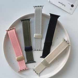 Könnte beinhalten: Fünf gewebte Apple Watch-Armbänder in den Farben Rosa, Olivgrün, Grau, Schwarz und Beige. Jedes Armband hat einen Metallverschluss. Die Armbänder werden auf einem weißen Teller präsentiert.