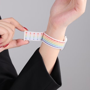 May include: White watch band with a rainbow stripe design. The band is made of a woven material and features a hook and loop closure. The rainbow colors include red, orange, yellow, green, blue, and purple.