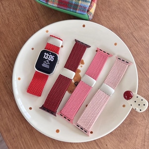 May include: An Apple Watch with a red braided band displays the time 13:05. Three additional braided watch bands in shades of red and pink are arranged on a white plate with brown polka dots. A small, decorative ceramic piece is also on the plate.