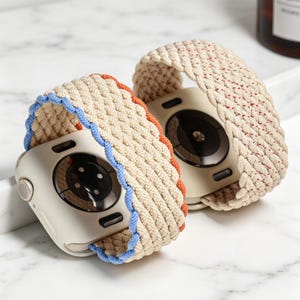May include: Two beige smart watch bands with braided designs. One band has blue and orange trim, the other has red trim. The watch cases are a light beige color. The bands are made of a woven material.
