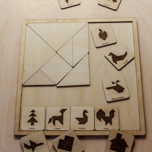 Może przedstawiać: Drewniana układanka tangram z kwadratową ramą i różnymi kształtami geometrycznymi. Zawiera mniejsze kwadratowe płytki z wytrawionymi ilustracjami zwierząt i przedmiotów, takich jak drzewo, łabędź i dom. Widoczne jest słowo "HELICOPTERE".