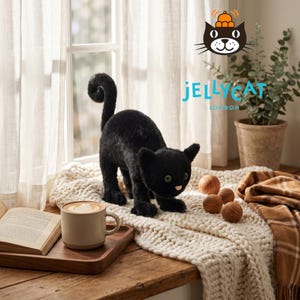 Può includere: Un giocattolo di peluche a forma di gatto nero con occhi verdi e coda arricciata è su una coperta di maglia color crema. Una tazza di caffè è su un vassoio di legno vicino a un libro aperto. Il logo Jellycat London è visibile.