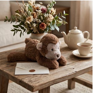 May include: A plush brown monkey toy with a light brown face and ears lies on a wooden table. A white teapot, teacup, and saucer are on the table. A bouquet of dried flowers sits in a vase. An envelope with a wax seal is also on the table.