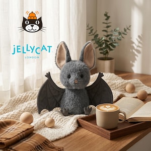 May include: A plush gray bat with large ears and outstretched wings sits on a cream-colored blanket. The bat has a soft, textured appearance. The image includes a wooden tray with a coffee cup, wooden spheres, and a book. The Jellycat London logo is visible.