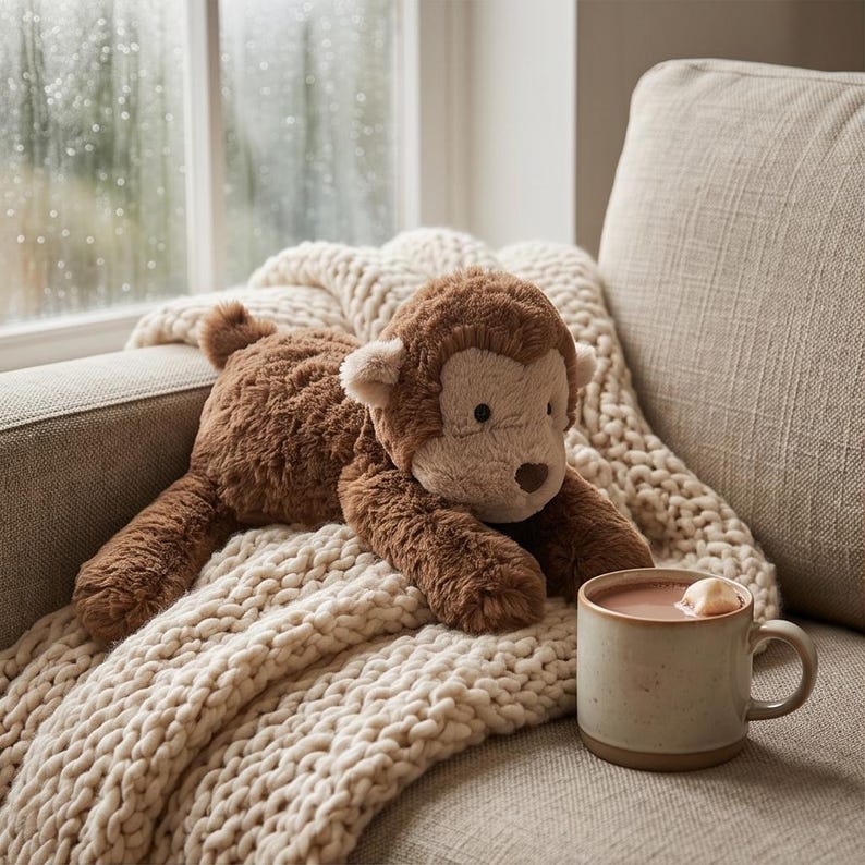 May include: A plush brown monkey toy with a light brown face and ears rests on a cream-colored knitted blanket. A mug of hot chocolate with a marshmallow sits nearby on a beige sofa. The scene is next to a window with raindrops.
