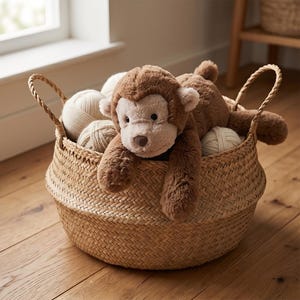 May include: A brown plush monkey toy rests in a woven basket filled with yarn balls. The basket has two handles and sits on a wooden floor. The monkey has a light brown face and dark brown fur.