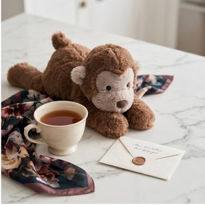 May include: A plush brown monkey toy with a light brown face and ears rests on a floral scarf. A cream-colored teacup filled with tea and a sealed envelope are nearby. The envelope has text.