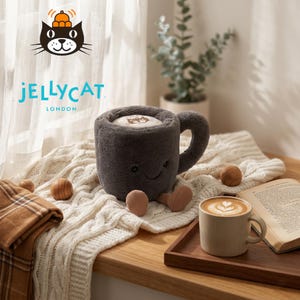 May include: A plush gray coffee mug toy with a smiling face, small beige feet, and a handle. The mug sits on a white knitted blanket with wooden balls and a small coffee cup. The Jellycat London logo is visible.