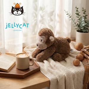 May include: A brown plush monkey toy with a light brown face and ears, lying on a cream-colored knitted blanket. A wooden tray holds a mug of coffee and an open book. The Jellycat logo is visible.