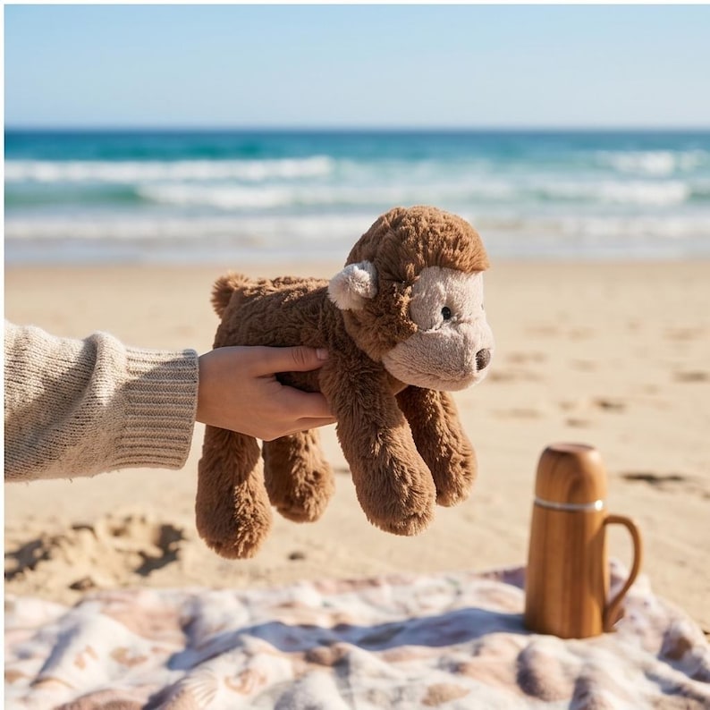 May include: A plush brown monkey with a light tan face and ears is held up against a beach backdrop. The monkey has a shaggy fur texture. A wooden thermos with a handle sits on a patterned blanket on the sand.