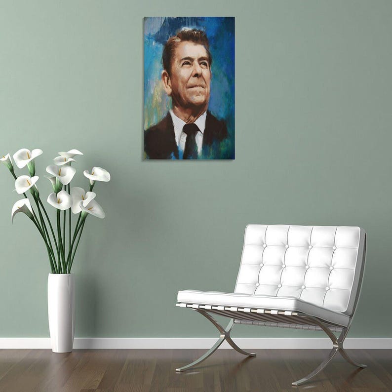 May include: A rectangular wall art print featuring a portrait of Ronald Reagan in a dark suit and tie, rendered in a painterly style with visible brushstrokes against a vibrant blue background, displayed in a room with a white chair and flowers.