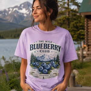 Puede incluir: Una camiseta de color morado claro con un estampado gráfico que incluye el texto THE WILD BLUEBERRY CLUB, ESTD 2026 y ALASKA MOUNTAINS PINES. El diseño muestra ilustraciones de arándanos, pinos y una cordillera con pequeñas estrellas.