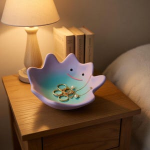 Peut inclure: Un vide-poches pastel en forme de personnage de dessin animé, contenant des bagues dorées et un collier de perles. Il est posé sur une table de chevet en bois, à côté d'une lampe et de livres.