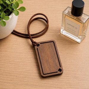 May include: A rectangular wooden pendant with a dark brown finish, featuring a recessed center and small spade symbols at the corners. The pendant is attached to a thin brown leather cord. A clear glass perfume bottle labeled SANTAL 33 is nearby.