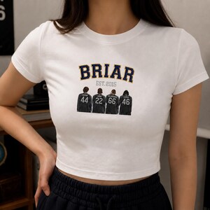 Puede incluir: Top corto blanco con la palabra "BRIAR" en azul marino y dorado. Debajo, cuatro figuras con chaquetas negras con los números 44, 22, 66 y 46. También está el texto "EST. 2015".