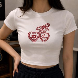 May include: White crop top with a red heart design. The design features two connected hearts with the number "22" in one heart and the name "John Logan" in the other. A red bow connects the hearts.