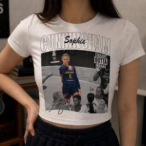 Puede incluir: Una camiseta blanca de manga corta con un estampado gráfico de la jugadora de baloncesto Sophie Cunningham. El diseño incluye el texto CUNNINGHAM, Sophie y STRAIGHT OUTTA INDIANA, junto con un collage en blanco y negro de escenas de juego.