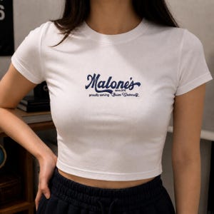 May include: White crop top with short sleeves. The shirt features the navy blue text "Malone's since 2015 proudly serving Briar University" in a cursive font. The shirt is made of a soft, comfortable material.