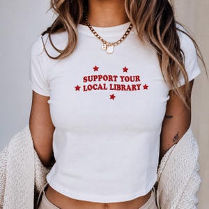 May include: A white short-sleeved t-shirt featuring red embroidered text that reads SUPPORT YOUR LOCAL LIBRARY, accented by five small red embroidered stars arranged around the lettering. The shirt has a crew neckline and a fitted silhouette.