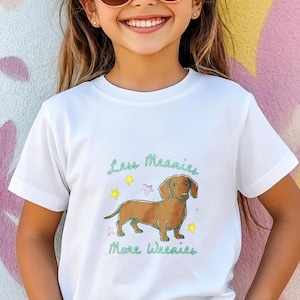 May include: A white short-sleeved t-shirt featuring a brown dachshund illustration in the center. The text "Less Meanies More Weenies" is printed in green above and below the dog, accompanied by small yellow and pink star graphics.