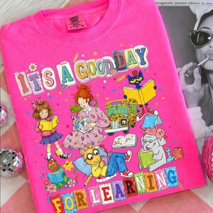 Puede incluir: Una camiseta de color rosa brillante con un estampado gr&aacute;fico colorido con el texto "IT'S A GOOD DAY FOR LEARNING". La ilustraci&oacute;n muestra a varios personajes de dibujos animados leyendo libros, incluyendo una ni&ntilde;a, una mujer, un gato y un elefante, junto a un autob&uacute;s escolar.