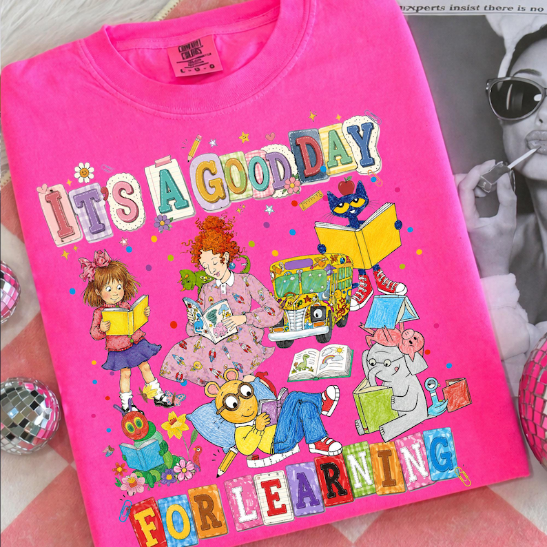 Puede incluir: Una camiseta de color rosa brillante con un estampado gr&aacute;fico colorido con el texto "IT'S A GOOD DAY FOR LEARNING". La ilustraci&oacute;n muestra a varios personajes de dibujos animados leyendo libros, incluyendo una ni&ntilde;a, una mujer, un gato y un elefante, junto a un autob&uacute;s escolar.