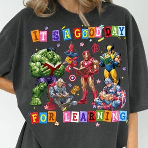 May include: A charcoal gray t-shirt featuring a graphic of Marvel superheroes reading books. The text reads IT'S A GOOD DAY FOR LEARNING. Characters include Hulk, Spider-Man, Iron Man, Wolverine, Thor, and Captain America with small flower icons.