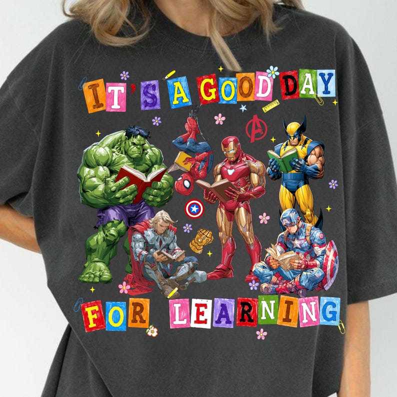 May include: A charcoal gray t-shirt featuring a graphic of Marvel superheroes reading books. The text reads IT'S A GOOD DAY FOR LEARNING. Characters include Hulk, Spider-Man, Iron Man, Wolverine, Thor, and Captain America with small flower icons.