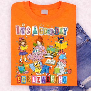 Puede incluir: Una camiseta naranja con una colorida ilustraci&oacute;n gr&aacute;fica de varios personajes de libros infantiles leyendo, incluyendo un autob&uacute;s escolar y el texto "IT'S A GOOD DAY FOR LEARNING" impreso en grandes letras de bloque multicolores al frente.