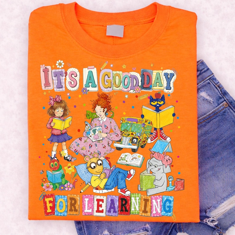 Puede incluir: Una camiseta naranja con una colorida ilustraci&oacute;n gr&aacute;fica de varios personajes de libros infantiles leyendo, incluyendo un autob&uacute;s escolar y el texto "IT'S A GOOD DAY FOR LEARNING" impreso en grandes letras de bloque multicolores al frente.