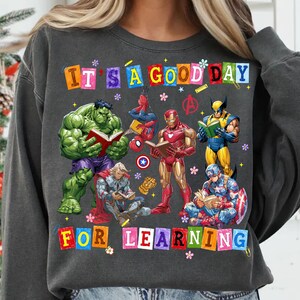May include: A charcoal gray crewneck sweatshirt featuring a graphic print of Marvel superheroes reading books. The text reads IT'S A GOOD DAY FOR LEARNING in colorful block letters. Characters include Hulk, Spider-Man, Iron Man, Wolverine, Thor, and Captain America.