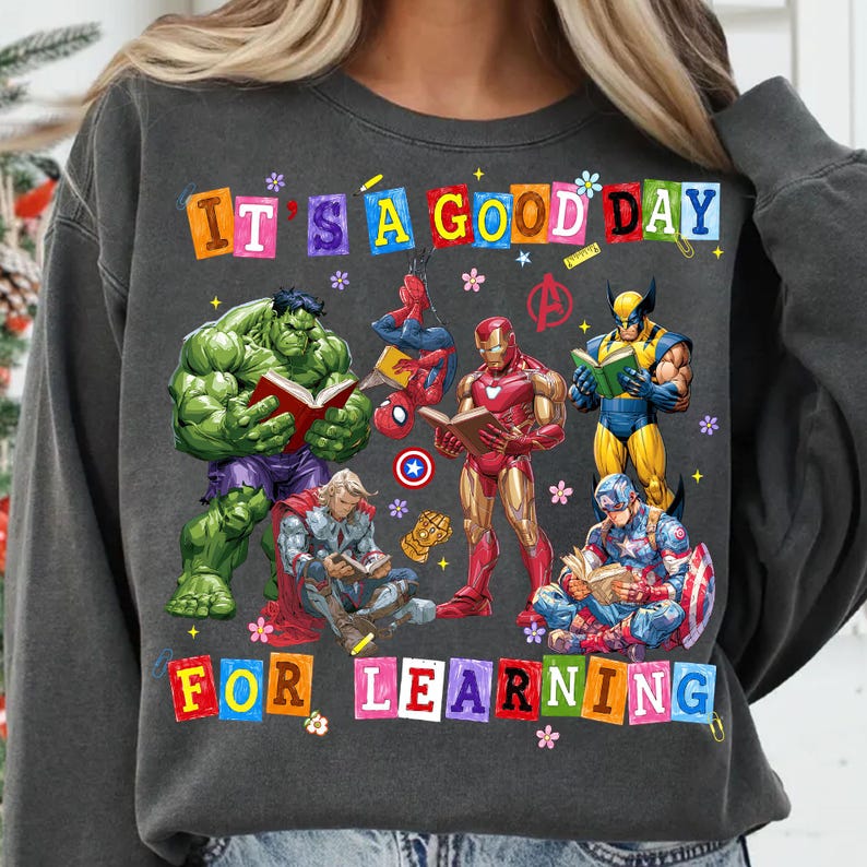 May include: A charcoal gray crewneck sweatshirt featuring a graphic print of Marvel superheroes reading books. The text reads IT'S A GOOD DAY FOR LEARNING in colorful block letters. Characters include Hulk, Spider-Man, Iron Man, Wolverine, Thor, and Captain America.