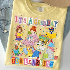 Puede incluir: Una camiseta de color amarillo p&aacute;lido con un estampado gr&aacute;fico colorido que incluye el texto IT'S A GOOD DAY FOR LEARNING. El dise&ntilde;o presenta ilustraciones de varios personajes de cuentos, un autob&uacute;s escolar y libros abiertos.