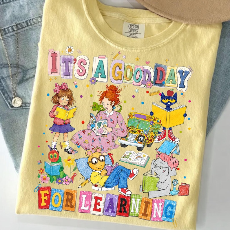 Puede incluir: Una camiseta de color amarillo p&aacute;lido con un estampado gr&aacute;fico colorido que incluye el texto IT'S A GOOD DAY FOR LEARNING. El dise&ntilde;o presenta ilustraciones de varios personajes de cuentos, un autob&uacute;s escolar y libros abiertos.