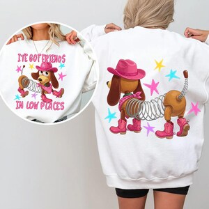 May include: A white sweatshirt featuring a graphic of a brown dachshund with a metal spring body, wearing a pink cowboy hat, pink bandana, and pink cowboy boots. The design includes colorful stars and the text I'VE GOT FRIENDS IN LOW PLACES.