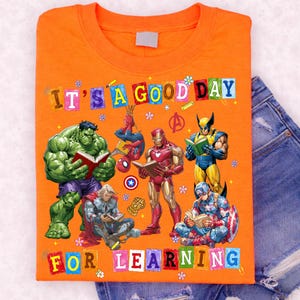 May include: An orange t-shirt featuring a graphic print of Marvel superheroes reading books, including Hulk, Spider-Man, Iron Man, Wolverine, Thor, and Captain America. The text reads IT'S A GOOD DAY FOR LEARNING in colorful block letters.