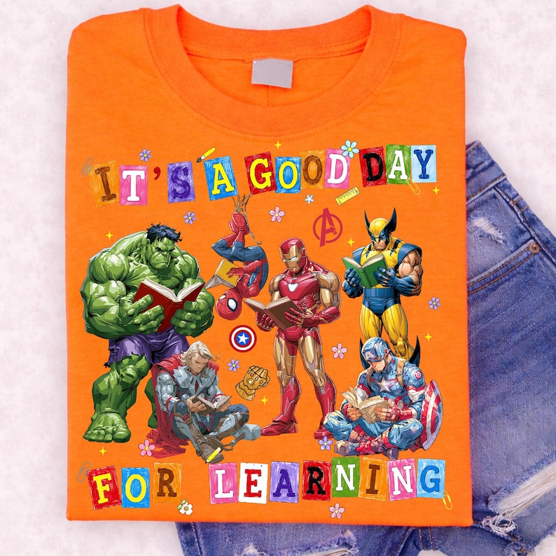 May include: An orange t-shirt featuring a graphic print of Marvel superheroes reading books, including Hulk, Spider-Man, Iron Man, Wolverine, Thor, and Captain America. The text reads IT'S A GOOD DAY FOR LEARNING in colorful block letters.