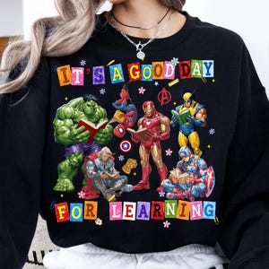 May include: A black crewneck sweatshirt featuring a graphic of Marvel superheroes including Hulk, Spider-Man, Iron Man, Wolverine, Thor, and Captain America reading books. The text reads IT'S A GOOD DAY FOR LEARNING in colorful block letters.