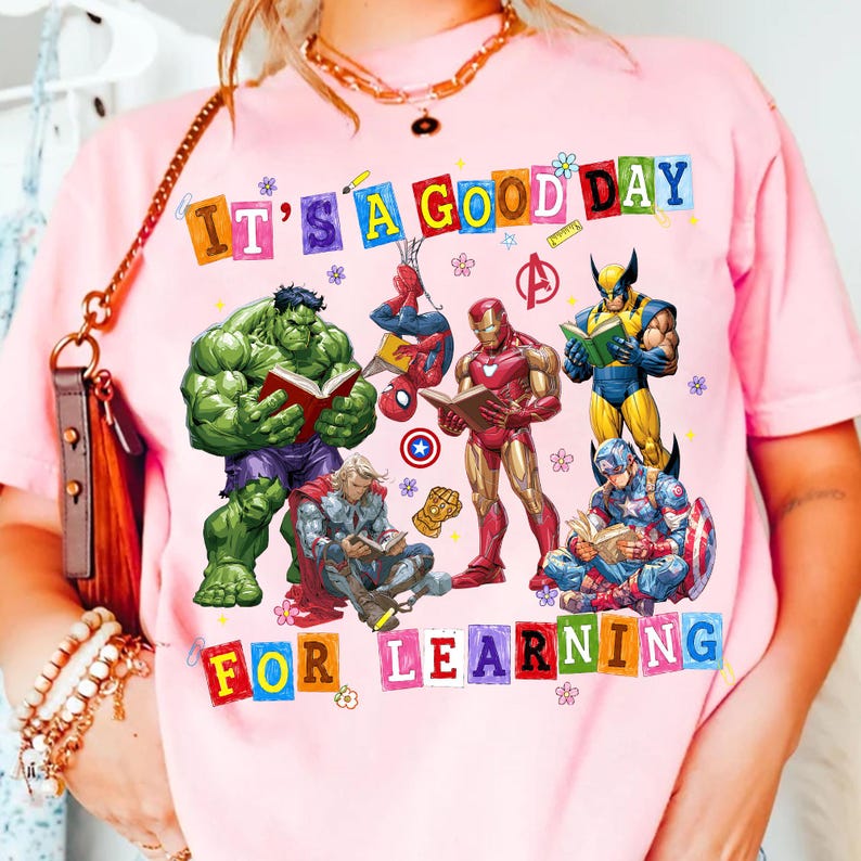 May include: A light pink t-shirt featuring a graphic of Marvel superheroes reading books. The text reads IT'S A GOOD DAY FOR LEARNING in colorful block letters. Characters include Hulk, Spider-Man, Iron Man, Wolverine, Thor, and Captain America.