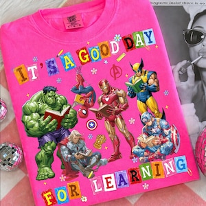 May include: A bright pink t-shirt featuring a graphic print of Marvel superheroes including Hulk, Spider-Man, Iron Man, Wolverine, Thor, and Captain America reading books. The text reads IT'S A GOOD DAY FOR LEARNING in colorful block letters.