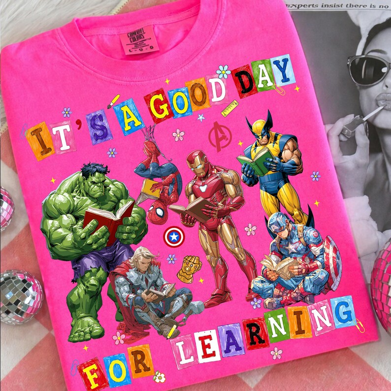 May include: A bright pink t-shirt featuring a graphic print of Marvel superheroes including Hulk, Spider-Man, Iron Man, Wolverine, Thor, and Captain America reading books. The text reads IT'S A GOOD DAY FOR LEARNING in colorful block letters.