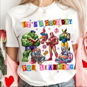 May include: A white t-shirt featuring a colorful graphic of Marvel superheroes reading books. The text "IT'S A GOOD DAY FOR LEARNING" is printed in multi-colored block letters. Characters include Hulk, Spider-Man, Iron Man, Wolverine, Thor, and Captain America.