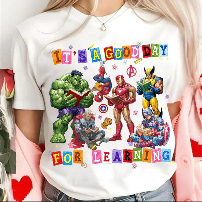 May include: A white t-shirt featuring a colorful graphic of Marvel superheroes reading books. The text "IT'S A GOOD DAY FOR LEARNING" is printed in multi-colored block letters. Characters include Hulk, Spider-Man, Iron Man, Wolverine, Thor, and Captain America.