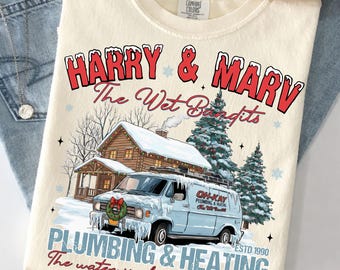 Puede incluir: Una camiseta color crema con un gráfico de una furgoneta azul claro frente a una cabaña nevada y pinos. El texto dice HARRY & MARV THE WET BANDITS, OH-KAY PLUMBING & HEATING ESTD 1990 y The water is always running.