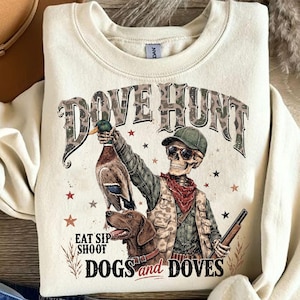 May include: A cream-colored crewneck sweatshirt featuring a graphic of a skeleton wearing a camouflage vest, a green cap, and sunglasses. The skeleton holds a duck and a shotgun, accompanied by a brown dog. Text reads DOVE HUNT and EAT SIP SHOOT.