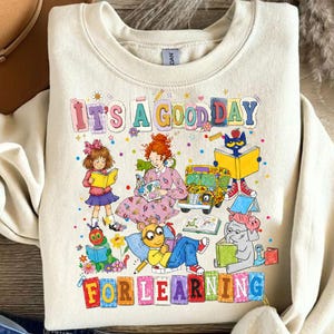 Puede incluir: Una sudadera de cuello redondo color crema con un estampado gr&aacute;fico colorido con el texto "IT'S A GOOD DAY FOR LEARNING". El dise&ntilde;o incluye ilustraciones de varios personajes de libros infantiles leyendo, un autob&uacute;s escolar amarillo y flores.