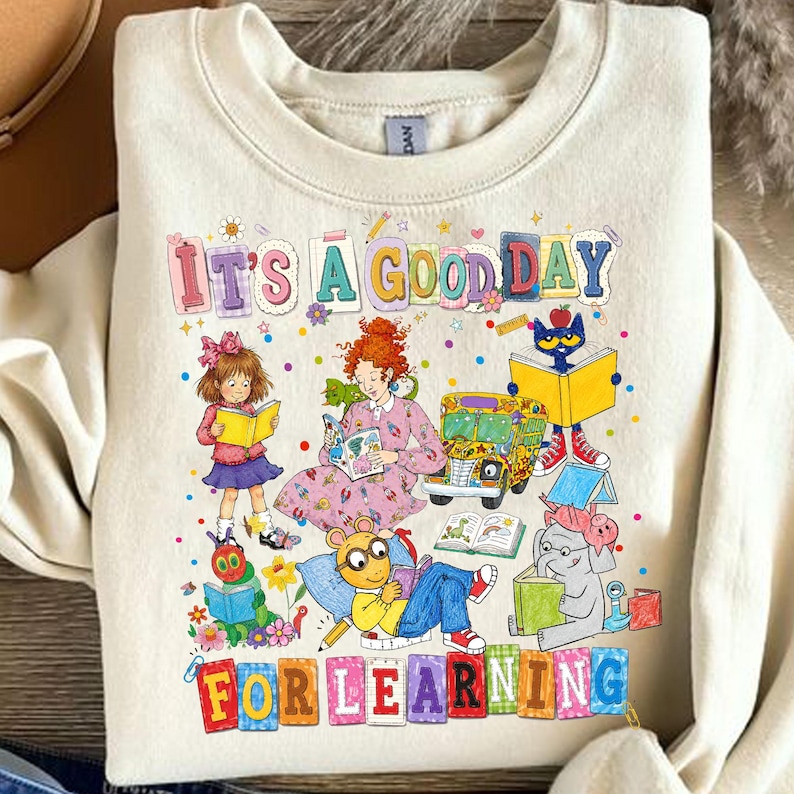 Puede incluir: Una sudadera de cuello redondo color crema con un estampado gr&aacute;fico colorido con el texto "IT'S A GOOD DAY FOR LEARNING". El dise&ntilde;o incluye ilustraciones de varios personajes de libros infantiles leyendo, un autob&uacute;s escolar amarillo y flores.