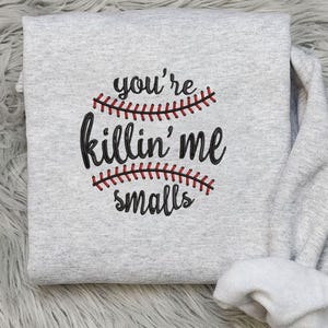 May include: A light gray sweatshirt with the phrase "you're killin' me smalls" in black script, with baseball stitching in red. The sweatshirt is folded on a fluffy, light-colored surface.